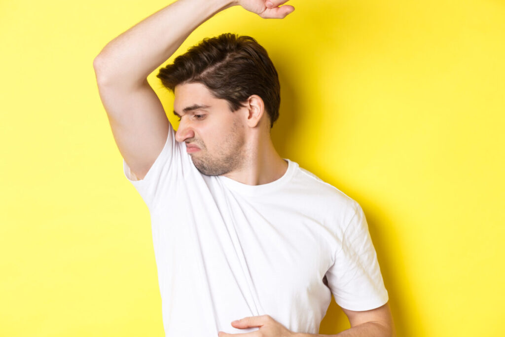 Man in sweat smelling his armpit, standing in white t-shirt and grimacing from stinky clothes