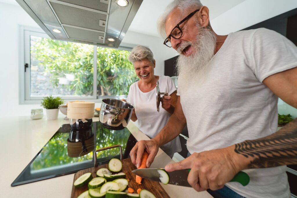 Happy senior couple having fun cooking together at home - Elderly people preparing health lunch in modern kitchen - Retired lifestyle family time and food nutrition concept
